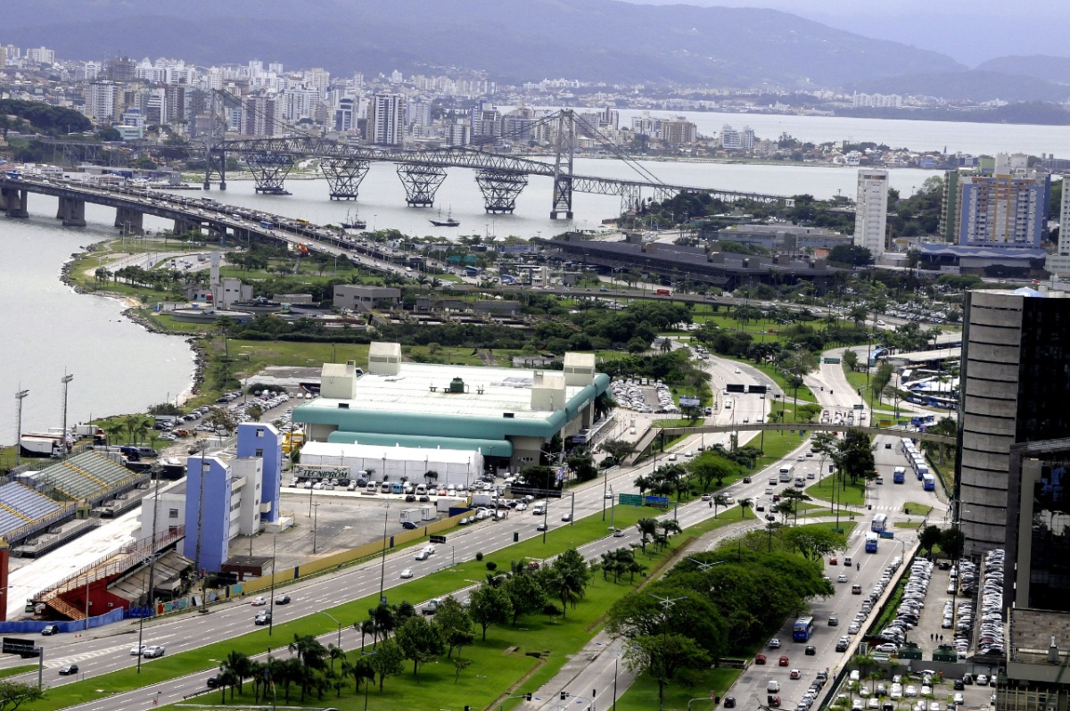 Foto colorida de uma paisagem urbana com um Centro de Eventos à esquerda várias vias asfaltadas ao centro e o mar com pontes ao fundo